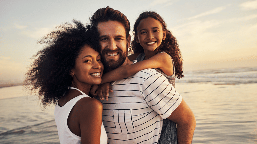Smiling family of three enjoying a day at the beach during sunset, with the child hugging both parents and the ocean in the background.