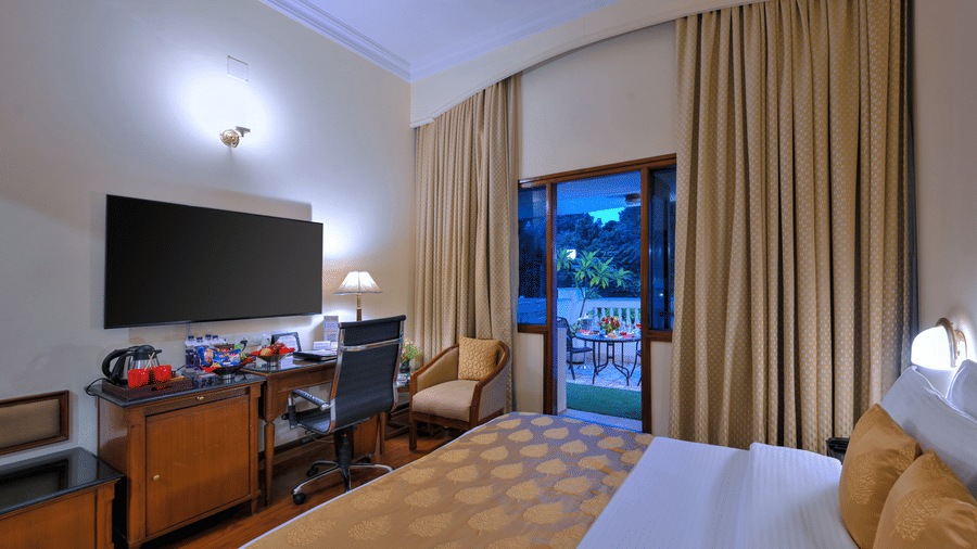 A room with a large bed, spreads, pillows, a TV, a chair, a table, and windows. | Clarks Varanasi