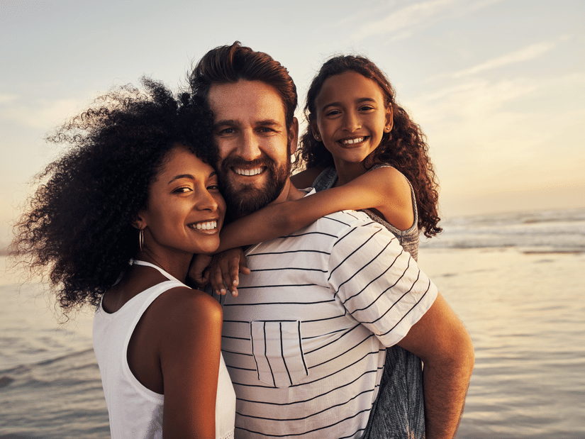 Smiling family of three enjoying a day at the beach during sunset, with the child hugging both parents and the ocean in the background.
