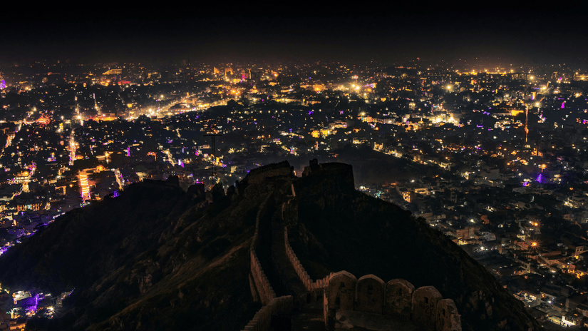The city of Jaipur glows with thousands of golden lights at night, viewed from the dark, high ridges of the fort.