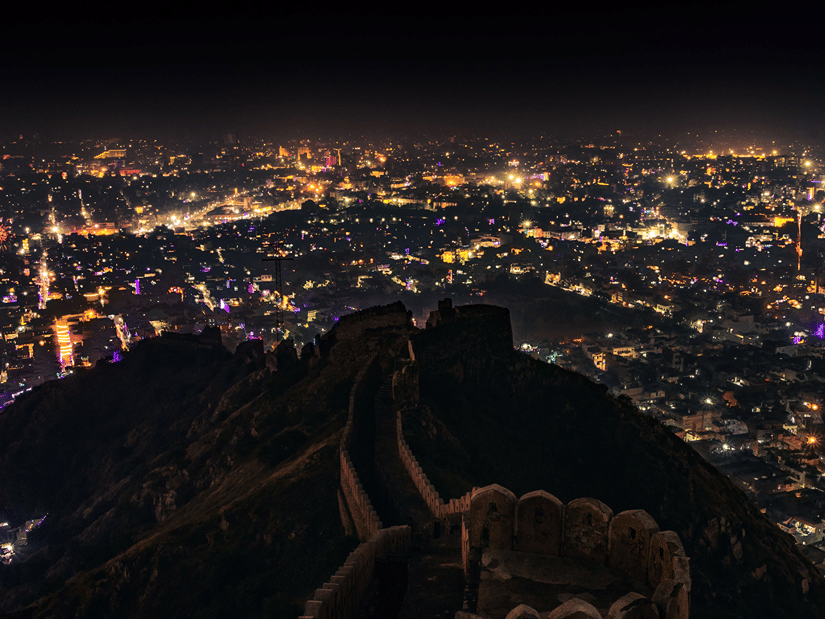 A panoramic night view of Jaipur, with thousands of illuminated buildings spread across the city and a dark hill in the foreground.