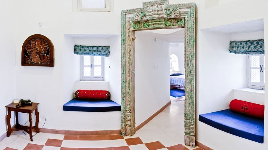 A room interior with a doorway, windows with cushioned seating, a small table, and a patterned floor at Tijara Fort-Palace - 19th Century, Alwar.