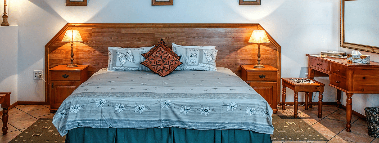 An image of a bedroom featuring a plush king size bed with blue sheets, bed side tables, lamps, a dressing table, a stool, and three photo frames hung on the wall