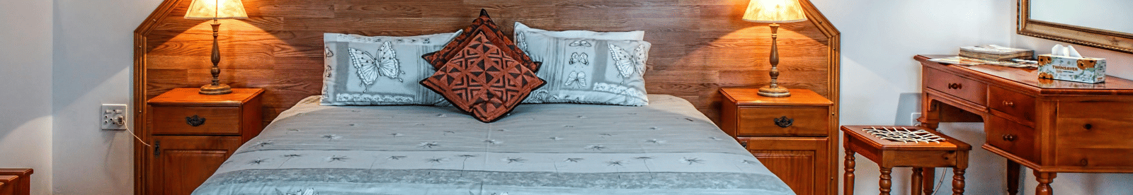 A room with a king size bed, bedside lamps, wall arts, and dressing mirror with a chair on the right.