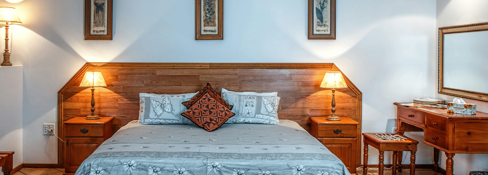 A cosy hotel bedroom with a large double bed, wooden headboard, two matching bedside tables and lamps, and three framed pictures.