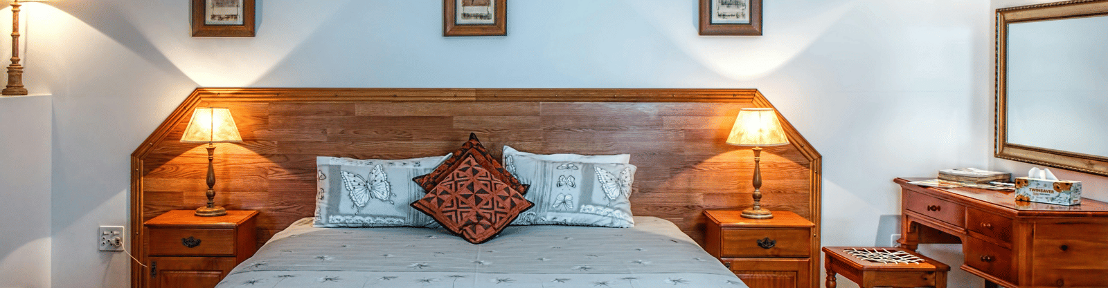 A cosy hotel bedroom with a large double bed, wooden headboard, two matching bedside tables and lamps, and three framed pictures.