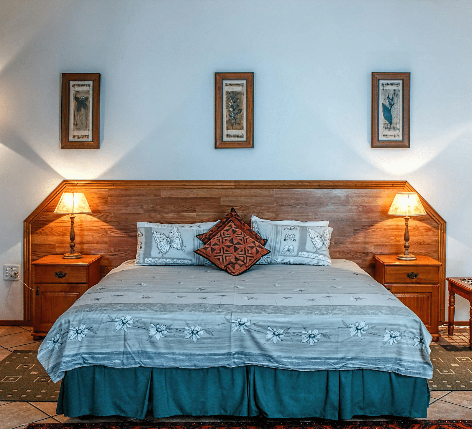 An image of a bedroom featuring a plush king size bed with blue sheets, bed side tables, lamps, a dressing table, a stool, and three photo frames hung on the wall