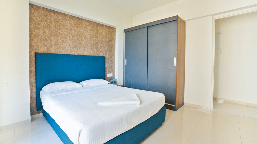 A minimalist bedroom with a blue upholstered bed, a grey wardrobe, and a textured gold feature wall behind the headboard at Apartel by Aarin, Oragadam.