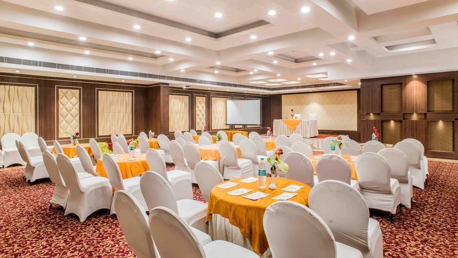 A banquet hall with round tables covered in orange cloths and white chairs at The Citi Residenci Hotel, Durgapur.
