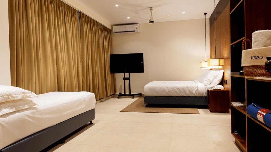 Hotel room at SUKAVI with 2 single beds, a wall-mounted television, and brown curtains covering the window.