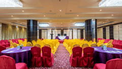 Symmetrical banquet hall with yellow-clad chairs, round tables with purple cloths, dark pillars, and bright ceiling lighting at Golden Tulip Kukas, Jaipur.