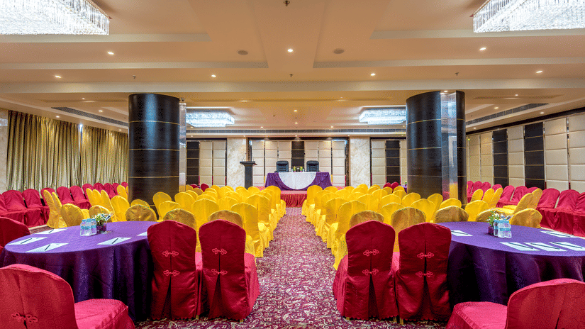 Symmetrical banquet hall with yellow-clad chairs, round tables with purple cloths, dark pillars, and bright ceiling lighting at Golden Tulip Kukas, Jaipur.