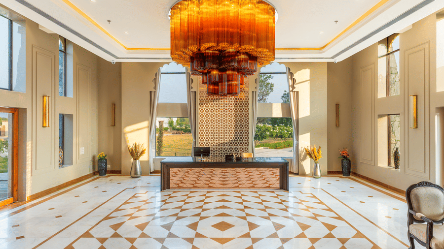The reception area at EsthereaRaj Leela, Ranakpur, featuring a dark wood desk, a large tiered orange chandelier, and checkkered marble floors.