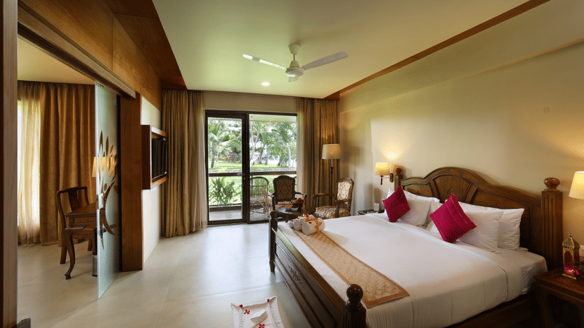 Lake View Suite at Uday Backwater Resort featuring a plush wooden bed with fresh linen and bed runner and bath towels rolled and tied with bows. In the background, there is a sitting space by the door to the balcony next to the lamp in the corner