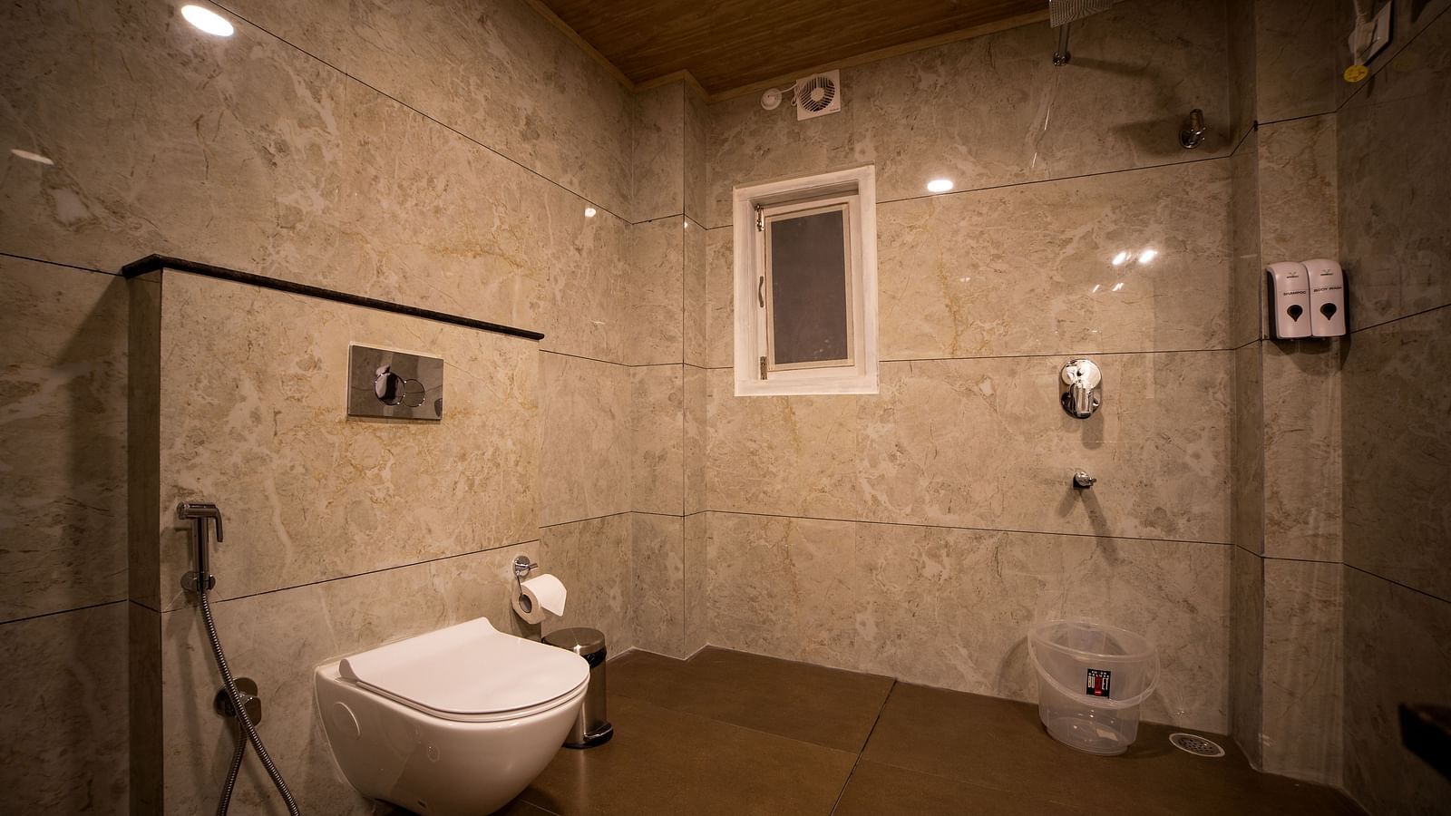 A bathroom with beige marble walls, a wall-mounted toilet, handheld bidet, shower area, and a small window for ventilation - Yashshree Pelling Chekhim Retreat.