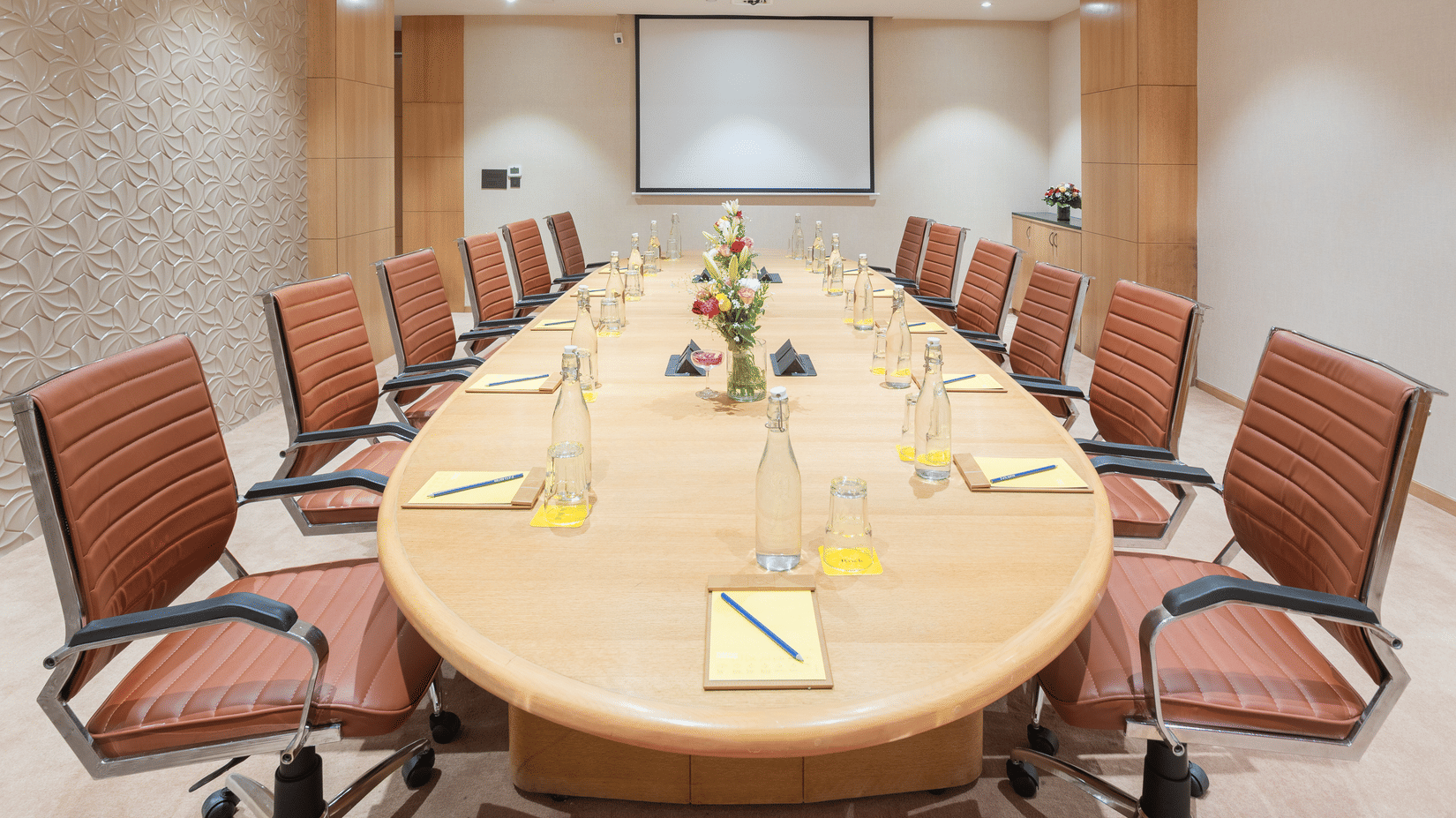 Board room at Golden Tulip Jaipur City Center with an oval table, leather chairs, notepads, water bottles, and a projector screen for meetings and presentations.