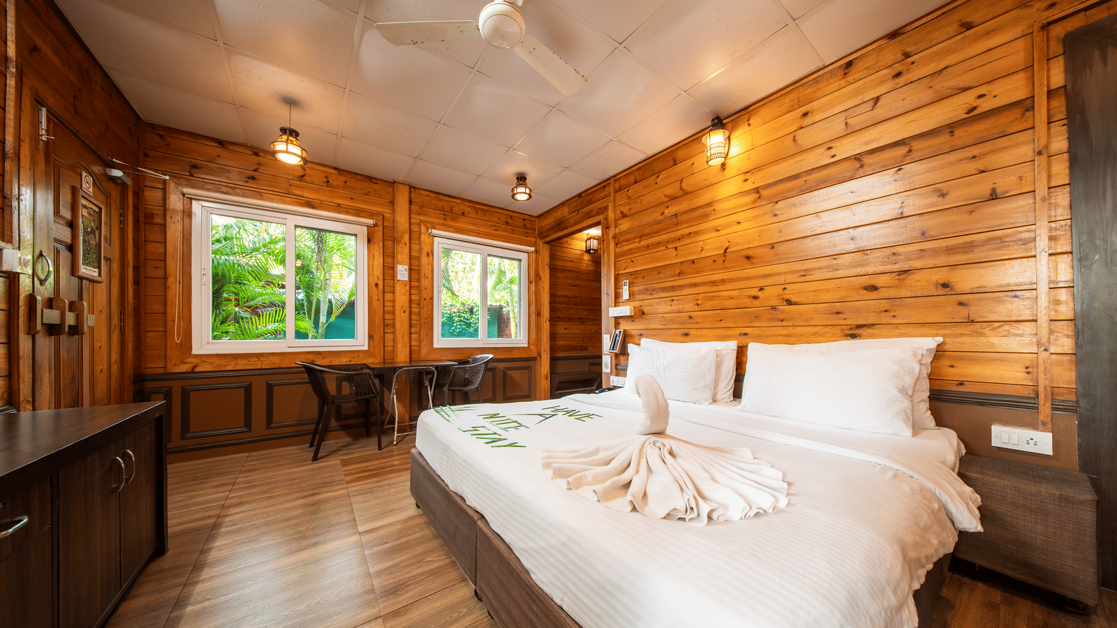 A corner angle view of room with wooden walls, floor, large windows, bed, and folded towels on the bed.