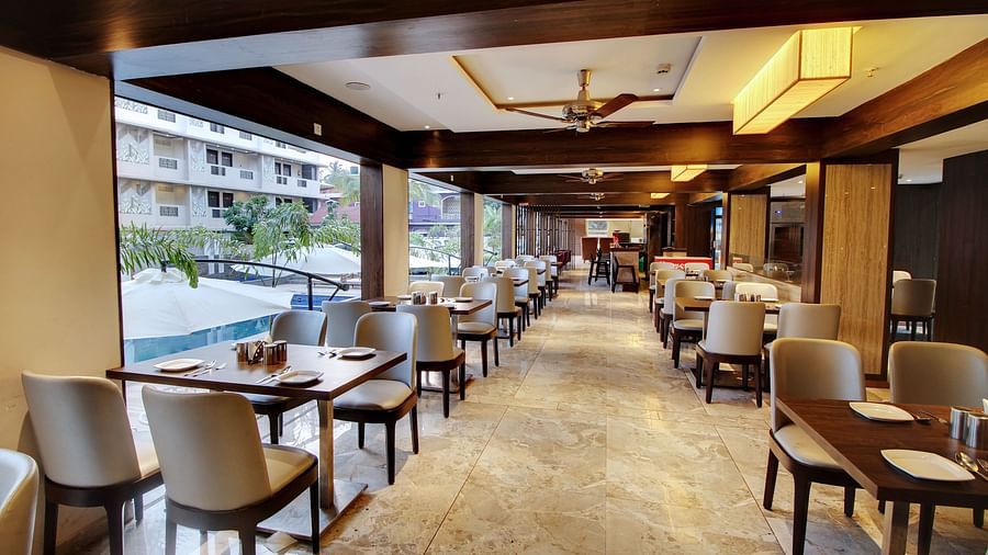 A hotel dining area with wooden tables and cream chairs, large windows providing natural light, and a view of the swimming pool outside
