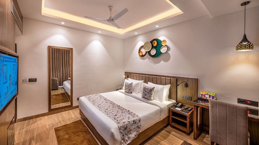 A hotel room with a king size bed, a flat-screen TV, and a work desk. It features a unique circular wall decoration, a ceiling fan, and a tray ceiling with lighting - Lords Eco Inn Dehradun.