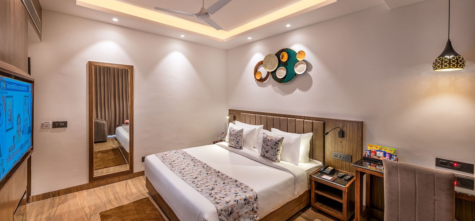 A hotel room with a king size bed, a flat-screen TV, and a work desk. It features a unique circular wall decoration, a ceiling fan, and a tray ceiling with lighting - Lords Eco Inn Dehradun.