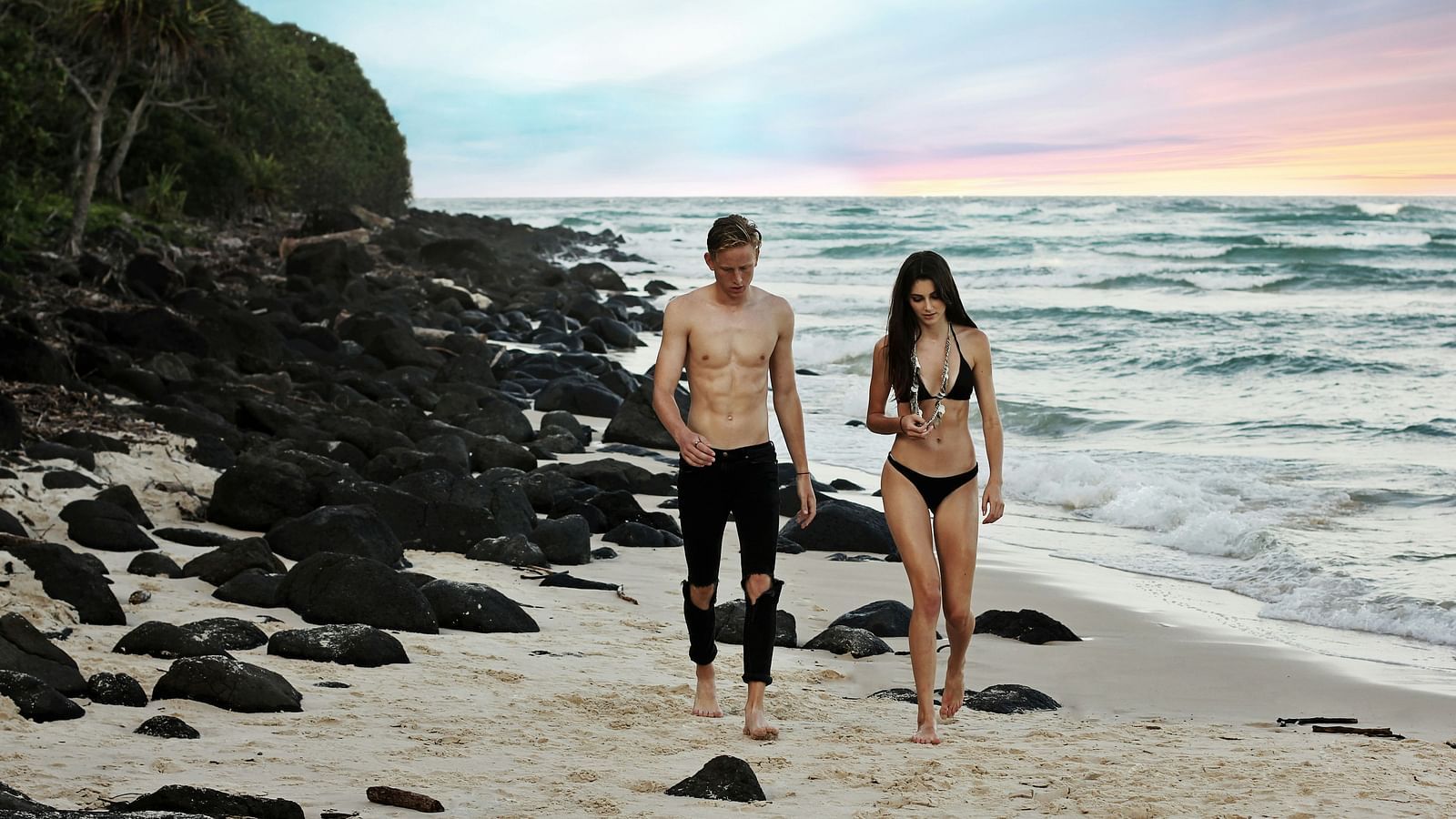 A couple walking in their swimsuits on a beach with rock formations on the left and the beach on the right.