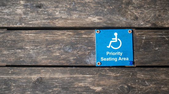 A sign attached to a weathered wooden surface with horizontal planks. The sign features a white international symbol for disability a person in a wheelchair above the words Priority Seating Area in white.