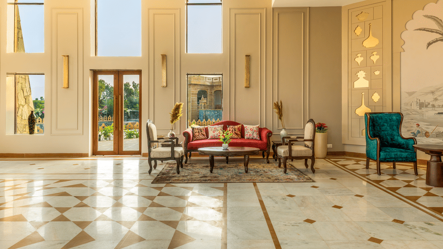 A view of the lobby featuring seating arrangements near glass doors that lead out to a sunlit garden area at EsthereaRaj Leela, Ranakpur.