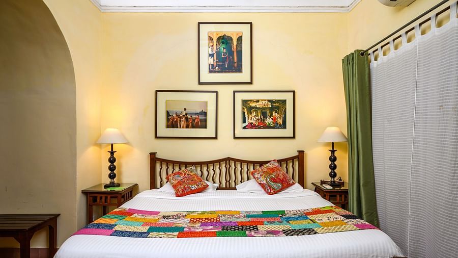 Room with bed, patterned runner and pillows, furniture including tables and lamps, framed artwork, curtains, and wall-mounted air conditioner - Tijara Fort-Palace - 19th Century, Alwar.