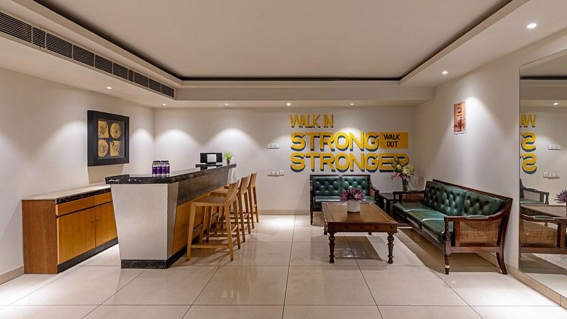 The reception area at Hotel Saket 27 is designed with a wooden desk with white ply finish paired with a green-upholstered sofa. The wooden structure and matching coffee table complement the overall warm and welcoming ambiance.
