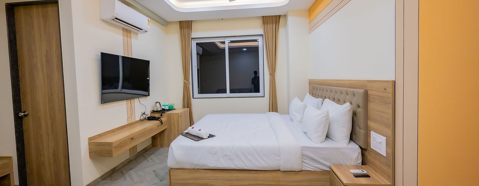 A well-lit modern hotel room with a comfortable double bed, wooden furniture, and a TV mounted on the wall.