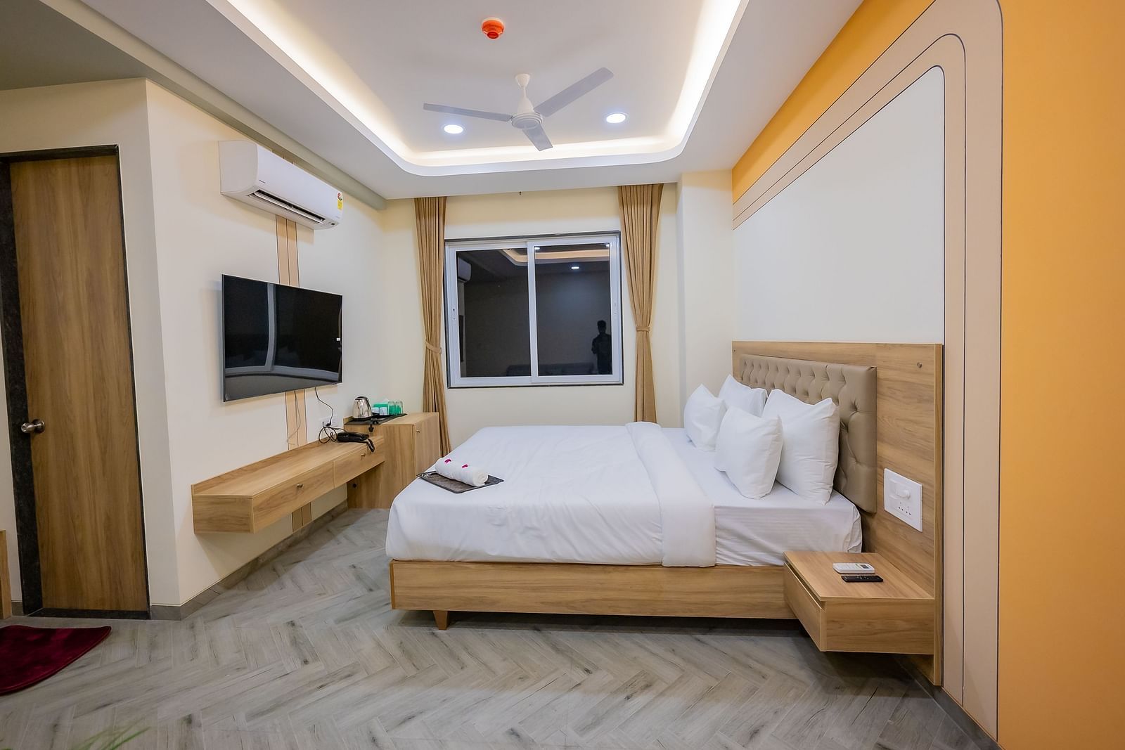 A well-lit modern hotel room with a comfortable double bed, wooden furniture, and a TV mounted on the wall.
