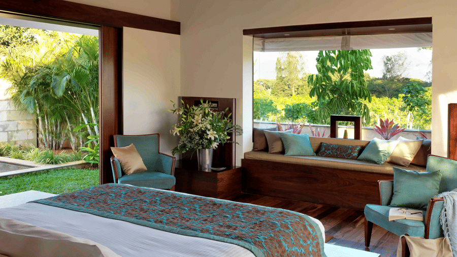Sunlit bedroom at The Serai Chikmagalur featuring a king bed with teal and brown bedding, seating area with cushions, large windows overlooking lush greenery.