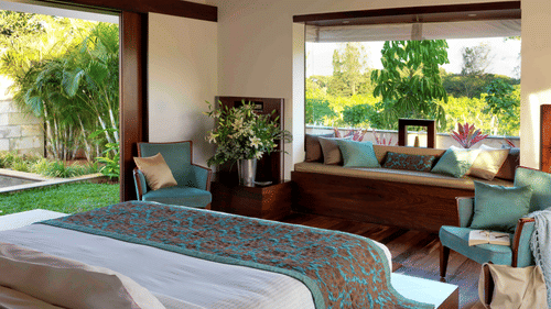 Sunlit bedroom at The Serai Chikmagalur featuring a king bed with teal and brown bedding, seating area with cushions, large windows overlooking lush greenery.