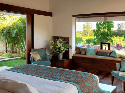 Sunlit bedroom at The Serai Chikmagalur featuring a king bed with teal and brown bedding, seating area with cushions, large windows overlooking lush greenery.