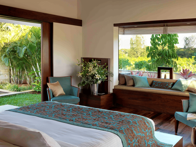 Sunlit bedroom at The Serai Chikmagalur featuring a king bed with teal and brown bedding, seating area with cushions, large windows overlooking lush greenery.