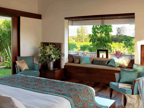 Sunlit bedroom at The Serai Chikmagalur featuring a king bed with teal and brown bedding, seating area with cushions, large windows overlooking lush greenery.