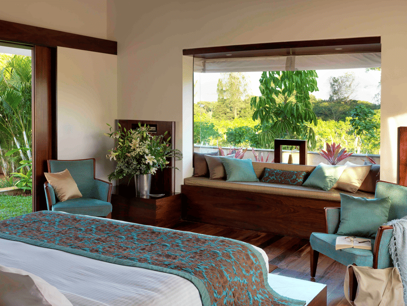 Sunlit bedroom at The Serai Chikmagalur featuring a king bed with teal and brown bedding, seating area with cushions, large windows overlooking lush greenery.
