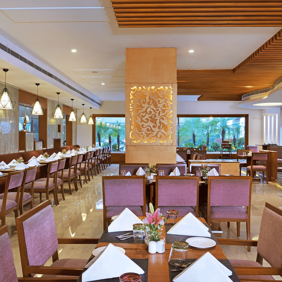 Coffee Shop restaurant at The Bristol Hotel, Gurgaon with banquette seating, a wooden slatted ceiling, pendant lights, and tables set with white napkins.