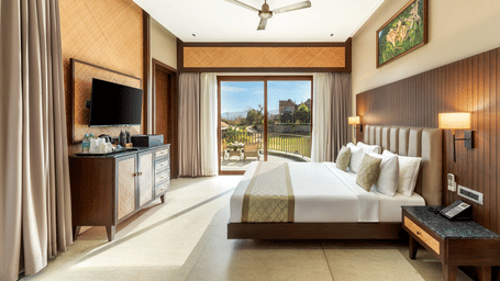 A bright, elegant hotel bedroom features a large bed, wall-mounted television, and glass doors to a balcony - Sajjan Bagh Resort and Spa, Kumbhalgarh