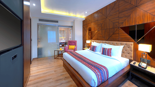An image of a bedroom with double size bed, bedside table next to it which has a lamp and a telephone on it, and at one corner there is a sofa and tea table - Golden Tulip, Agra