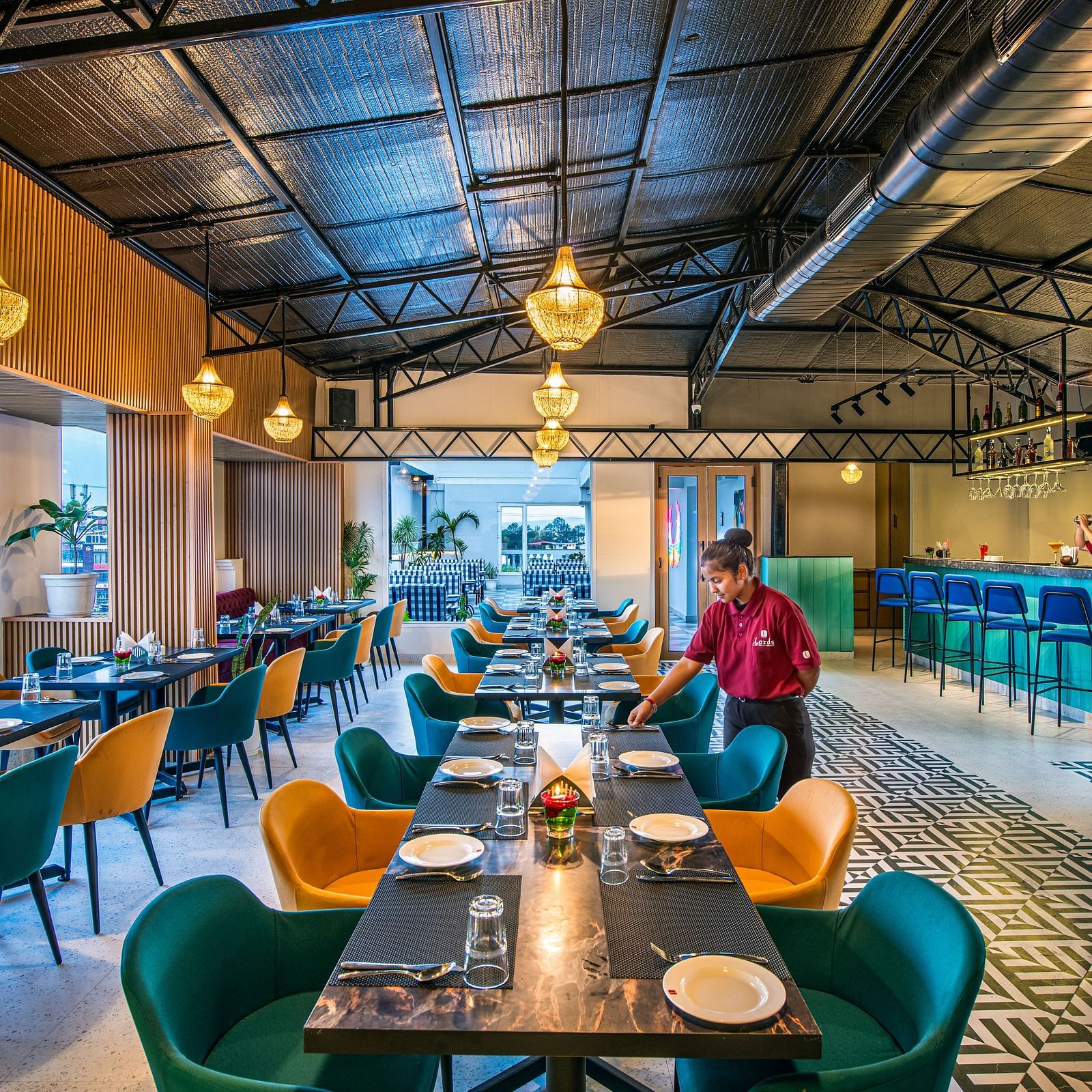 Restaurant with rows of tables set for dining, surrounded by colourful upholstered chairs, and a staff member visible. It features a high, exposed ceiling, unique pendant lighting, and a patterned tile floor - Lords Eco Inn Dehradun.