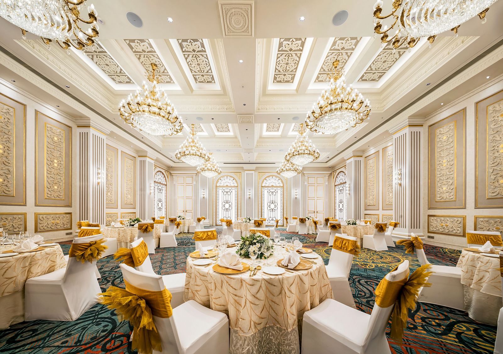  A lavish banquet room at Grand Chennai by GRT Hotels, showcasing numerous round tables dressed in elegant drapes, surrounded by white and gold chairs, all under the ornate light of sparkling chandeliers.