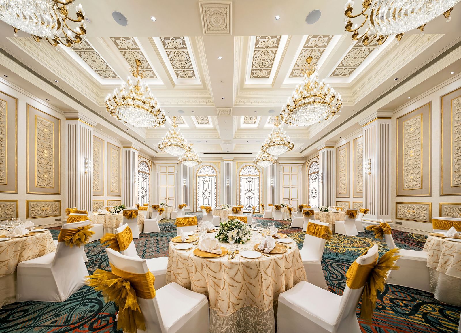  A lavish banquet room at Grand Chennai by GRT Hotels, showcasing numerous round tables dressed in elegant drapes, surrounded by white and gold chairs, all under the ornate light of sparkling chandeliers.