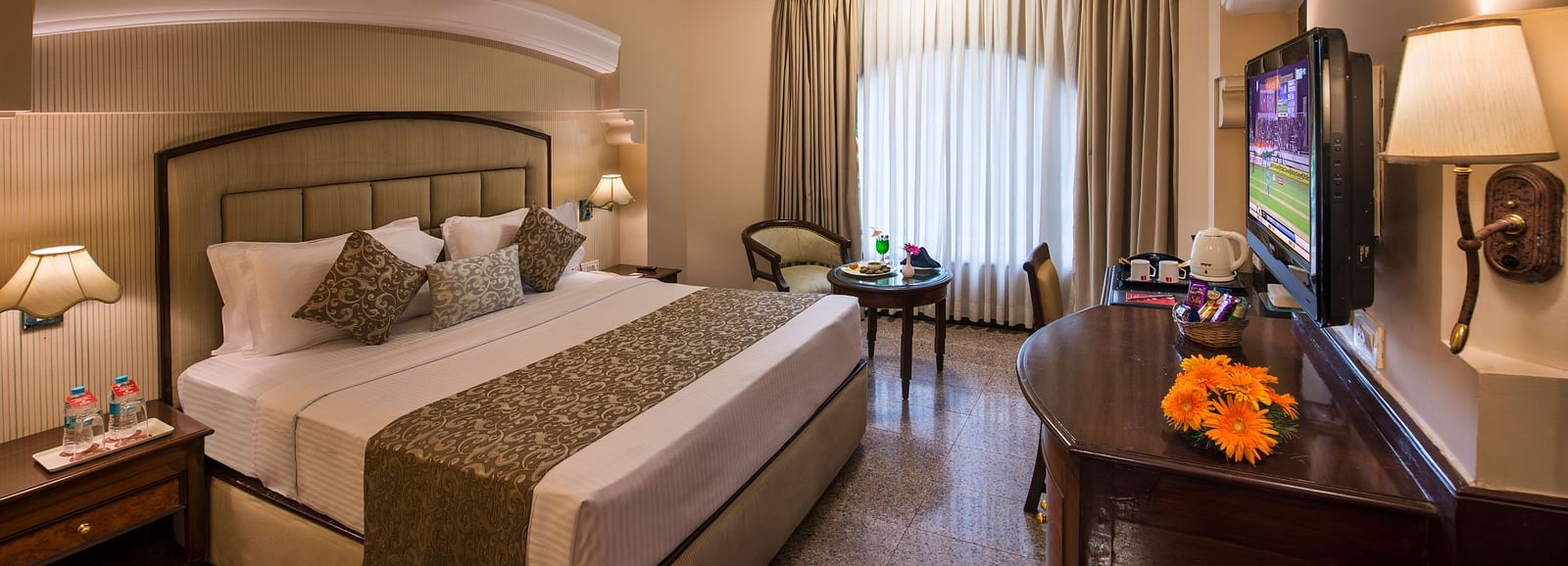 Hotel room with a king-sized bed, a decorative lamp, and a view of the outside through a window - Lords Inn Vadodara