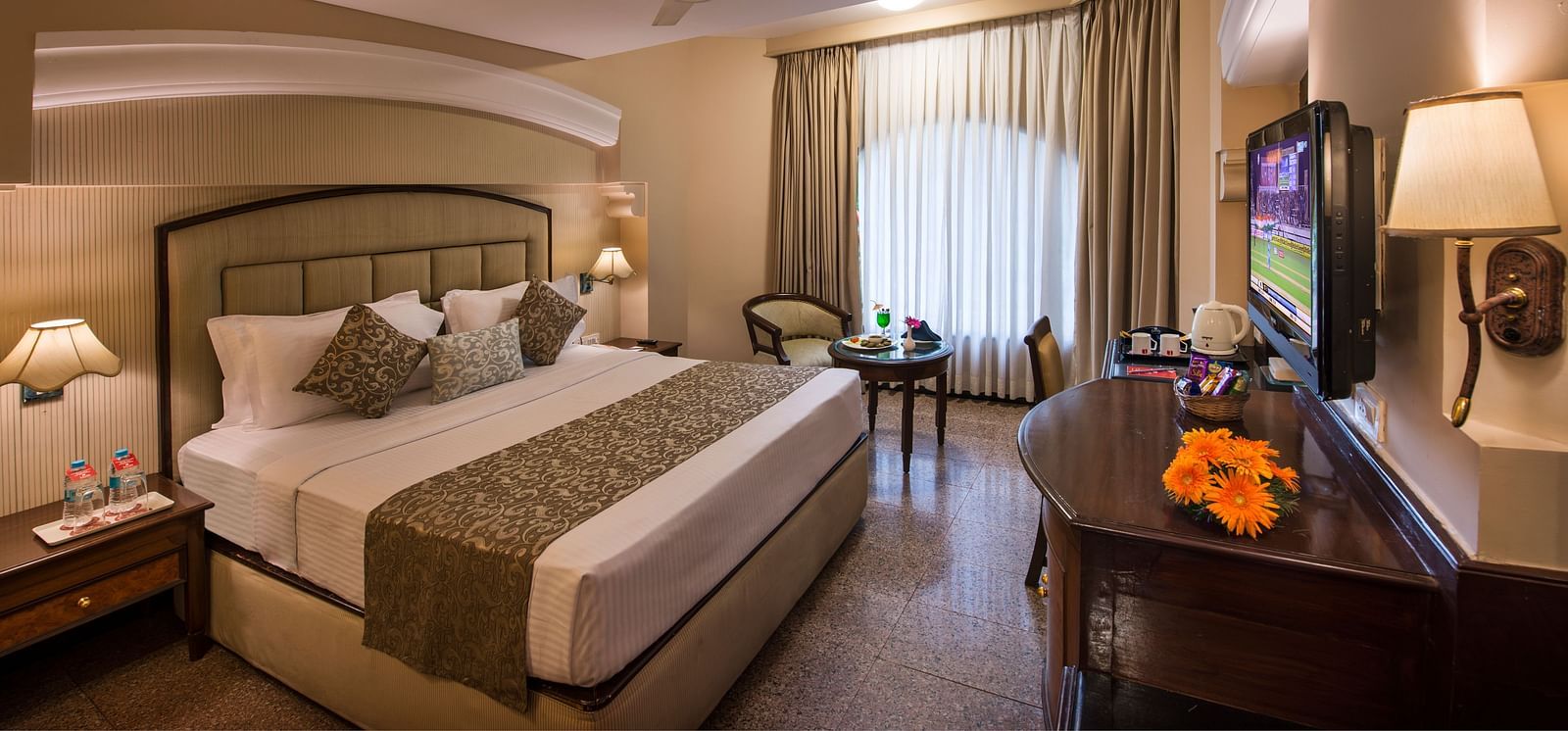 Hotel room with a king-sized bed, a decorative lamp, and a view of the outside through a window - Lords Inn Vadodara