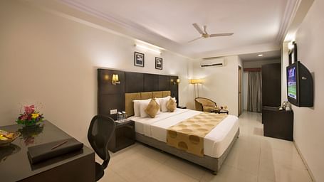 A hotel bedroom with a double bed, dark headboard, a desk and chair, a television, and a seating area with an armchair - Lords Eco Inn Dahej