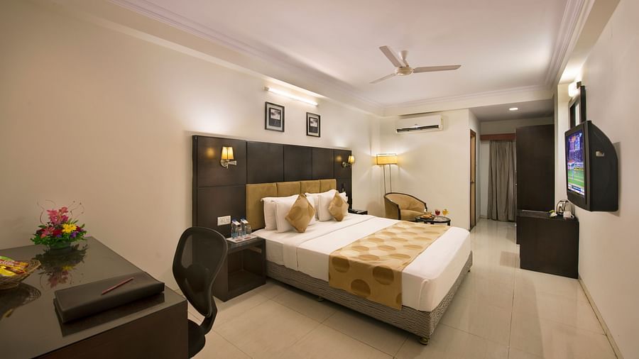 A hotel bedroom with a double bed, dark headboard, a desk and chair, a television, and a seating area with an armchair - Lords Eco Inn Dahej