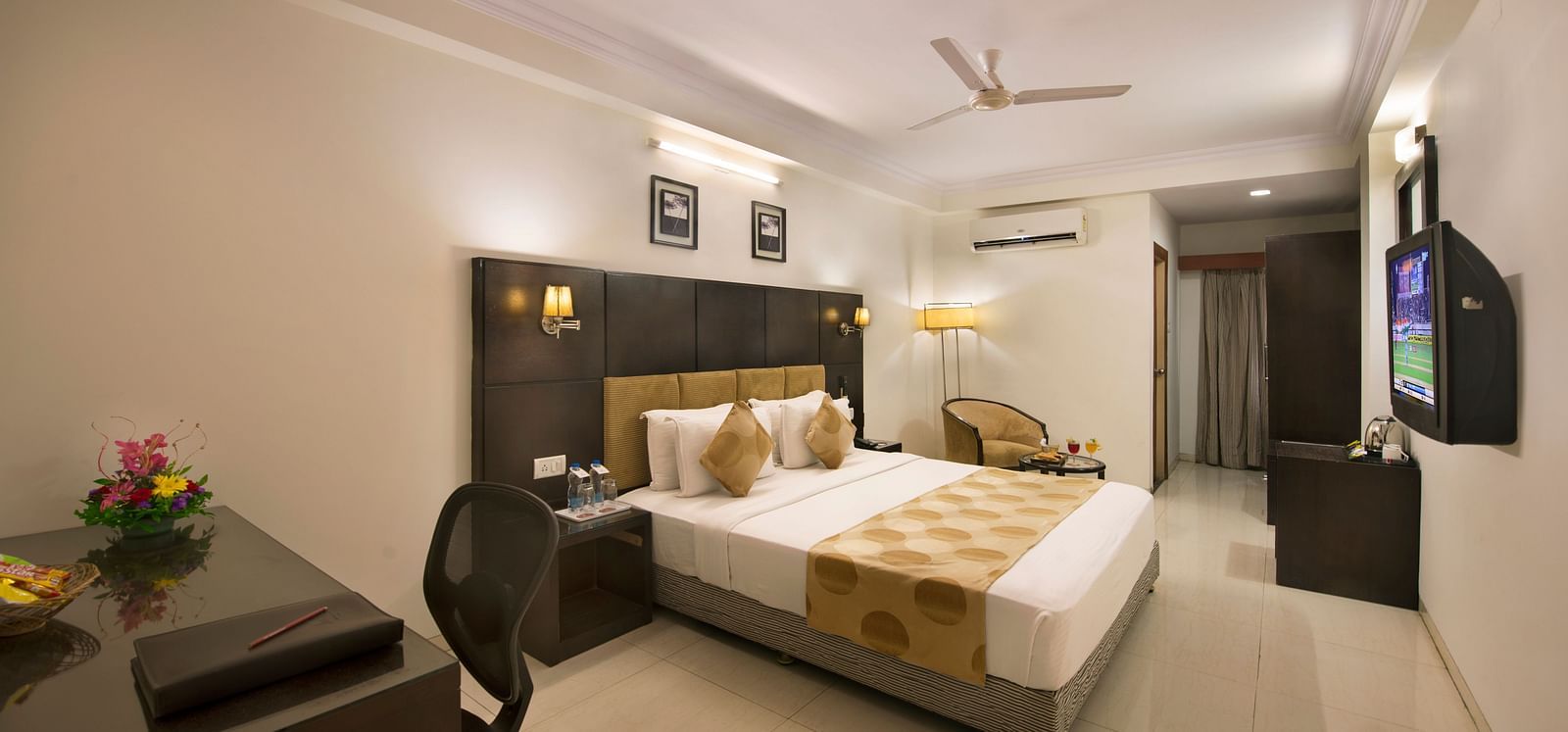 A hotel bedroom with a double bed, dark headboard, a desk and chair, a television, and a seating area with an armchair - Lords Eco Inn Dahej