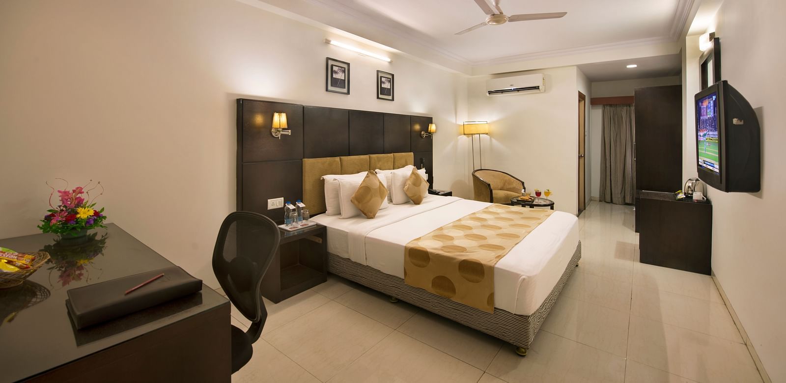 A hotel bedroom with a double bed, dark headboard, a desk and chair, a television, and a seating area with an armchair - Lords Eco Inn Dahej