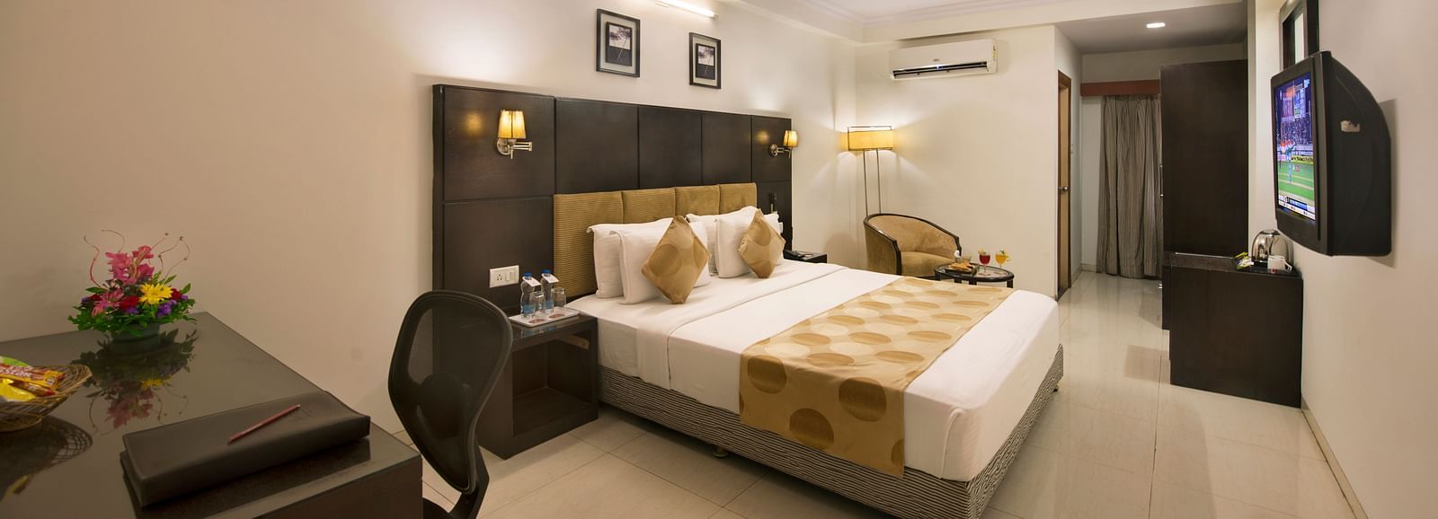 A hotel bedroom with a double bed, dark headboard, a desk and chair, a television, and a seating area with an armchair - Lords Eco Inn Dahej
